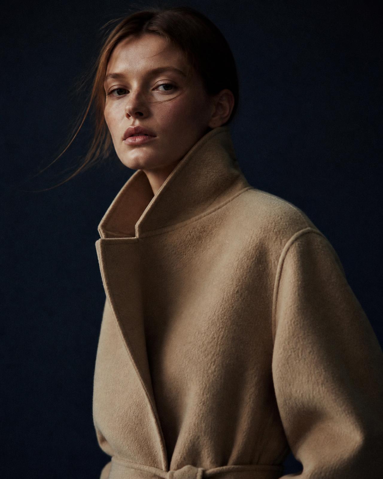 Editorial campaign frame of a camel wool coat against a navy studio backdrop